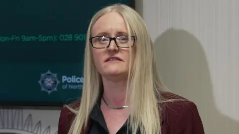 Geraldine Hanna has her mouth slightly open as she speaks at a press conference. She has long blonde hair, and silver necklace. She is wearing a wine coloured suit jacket and a black shirt.