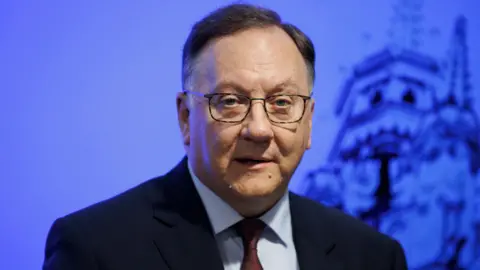 PA Media The Reverend Trevor Gribben speaks to media. He is an older man with short, dark, greying hair. He is wearing a pair of rectangular glasses and a dark suit jacket, a blue collared shirt and a red tie. He is stood in front of a blue screen.