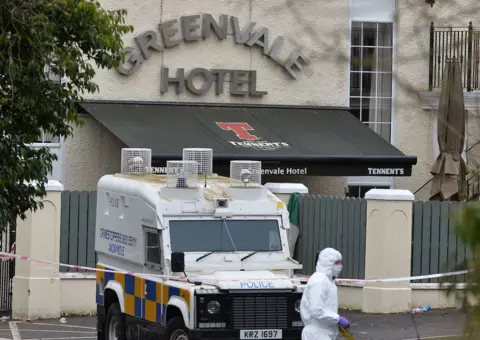 Pacemaker A Police Service of Northern Ireland is parked behind red and white police tape in front of the entrance to the Greenvale Hotel. Behind the land rover is a grey fence, which is in front of an awning under a sign in silver letters saying 'Greenvale Hotel'. In the foreground of the picture, in front of the police land rover, is a forensic officer wearing white protective gear from head to toe and purple gloves