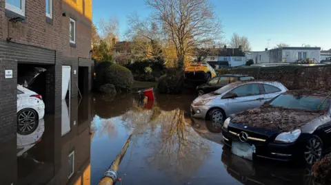Three cars are parked outside residential homes submerged in flood water.