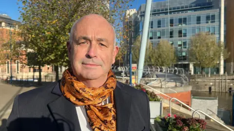 Phil Smith faces the camera. He is wearing a dark grey blaze with a white shirt and a scarf with an orange and black paisley-style pattern. Behind him are several tall, modern buildings which are part of the business district around Bristol Temple Meads station