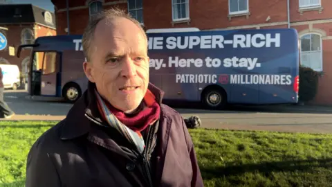 Stephen Kinsella faces the camera wearing a black jacket and a colourful scarf tucked inside it at the neck. He is a middle-aged man with wispy hair and behind him is a grassy area