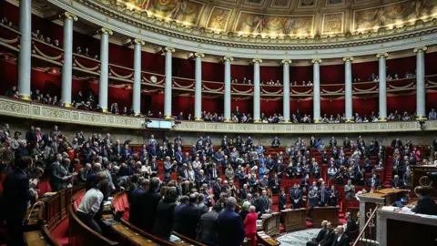 ALAIN JOCARD/AFP via Getty Images A wide shot inside the parliament building, you can see many people, all standing. The room is very beautiful, with intricate gold and pillars, and you can see the ornately painted ceiling.