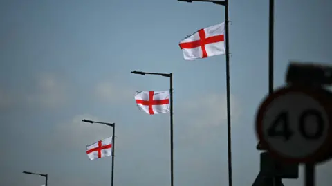 Getty Images Three street lamps with St George's crosses hanging near the tops against a blue sky with white clounds. In the foreground, a 40mph speed limit sign.