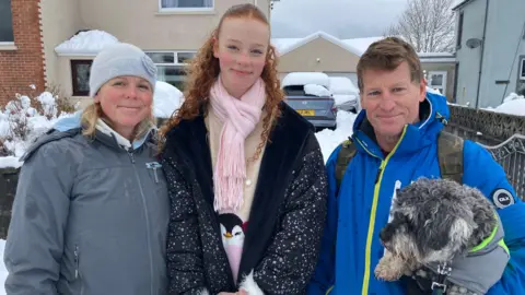 Helen Mcloughlin, Ela Mcloughlin, 14, Conan Blake and dog Madog, wearing winter gear, pose and smile outside a snow-covered home and car.