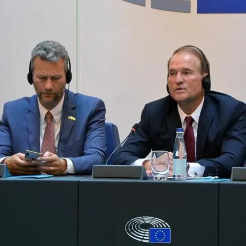 Facebook Two men sitting at a table at a meeting at the European Parliament. The grey-haired man on the left has grey stubble and is wearing a blue suit, pink shirt and a dark pink tie and is looking at his phone while wearing headphones. The man on the right is wearing a navy blue suit with a light pink shirt and dark red tie and wearing headphones. In front of them is a table with a glass, a bottle or water and a microphone 