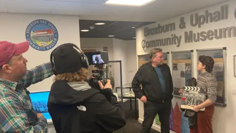 Caton-Jones talks to a young person holding a clapperboard with his name on it while two others point a camera at him and record the exchange. A sign saying Broxburn and Uphall Community Museum is on the wall above them.