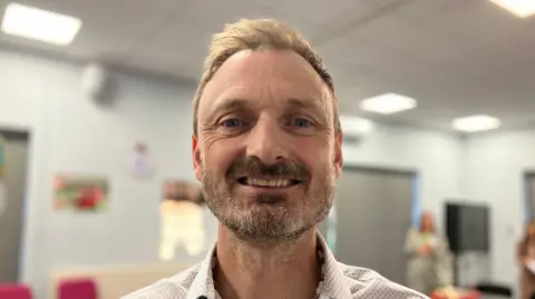 Shariqua Ahmed/BBC Matt Oliver looks directly at the camera smiling. He is wearing a white-patterned shirt and has short brown hair and a beard. 