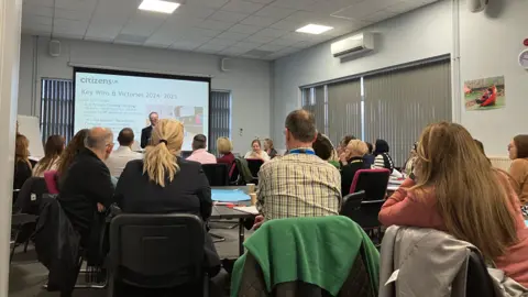 Shariqua Ahmed/BBC A group of people sit at tables facing the projector screen and a man speaking in front of it. The photograph is taken from the back of the room. 