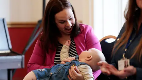 Getty Images Kate Forbes, in pink blazer and black and white stripe top, sits in a mother and baby group, holding her baby daughter in her lap and smiling down at her.