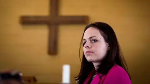 Getty Images Kate Forbes looks seriously off to the side, standing in a church, a wooden cross behind her and a white candle below it. She wears a pink blazer.