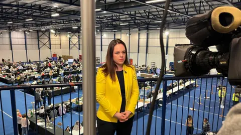 Getty Images Kate Forbes, in a bright yellow blazer stands on a gantry in front of a TV camera, an electoral count going on at tables in the room below her.