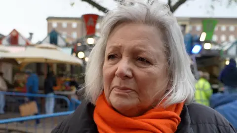 Qays Najm/BBC Sarah Langshaw is looking at the camera. She has shoulder-length grey hair and is wearing an orange scarf and a black coat. She is standing in front of a market.