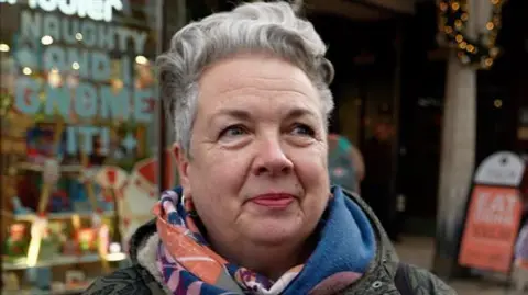 Qays Najm/BBC Emma Rope has short grey hair and is wearing a patterned scarf and a green coat. She is standing in front of a shop. 