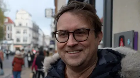 Qays Najm/BBC David Jackson has short brown hair and is wearing black-framed glasses. He is wearing a navy coat and is standing outside a building. He has a wide grin.