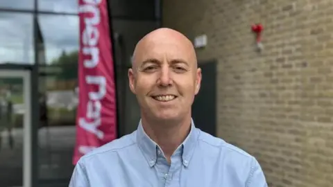 Jon Burrows is bald and is wearing a light blue shirt open at the neck and is smiling to the camera