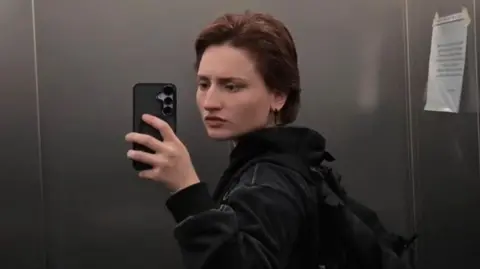 Aishat Baimuradova/Instagram Aishat taking a mirror selfie inside an elevator. She is wearing a dark hoodie and carrying a black backpack