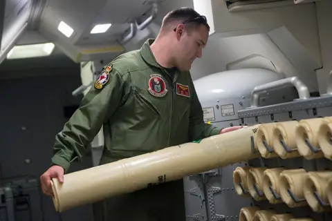 Getty Images A US naval officer loads a sonobuoy onto a rack for use in the search for Flight MH370. He wears an army green jumpsuit and has a buzz-cut hair style. The sonobuoy he is loading is cylindrical and beige in colour. He appears to be on a aeroplane. 