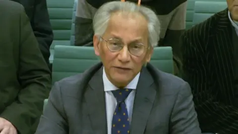 House of Commons Samir Shah giving evidence at the committee - he is sitting on a green chair, with other people behind him. He has grey hair and thin-rimmed glasses and is wearing a grey suit jacket with a striped shirt and blue spotted tie 