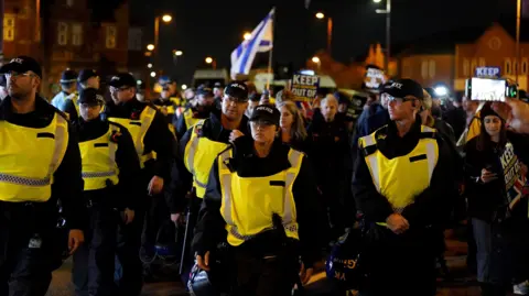 PA Media Numerous police officers in caps and hi-vis tabards stand in front of protesters carrying placards and Israeli flags on a city street at night. Some of the placards read "Keep antisemitism out of football". 