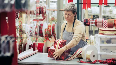 Getty Images A woman wearing an apron stands behind a counter wrapping Christmas presents with a thick red and gold ribbon. Reels of ribbons are seen behind her.