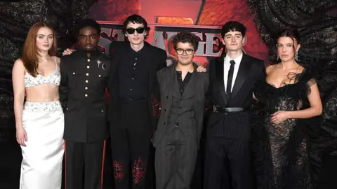 Getty Images The cast of Stranger Things stand in mostly dark clothing in front of the show's branding at the show's Dubai premiere. Sadie Sink, Caleb McLaughlin, Finn Wolfhard, Gaten Matarazzo, Noah Schnapp and Millie Bobby Brown are all in attendance.