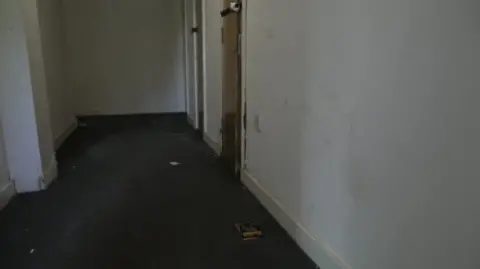 The image shows an empty corridor in one of the blocks, with litter strewn on the floor.