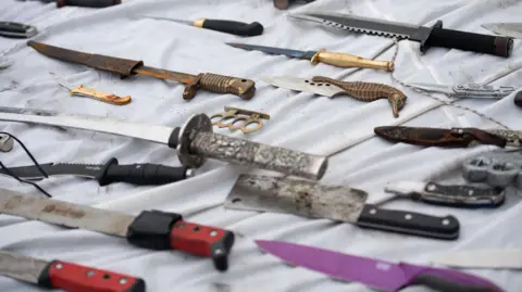 PA Media Knives and other weapons laid on a white sheet that have been deposited into weapon surrender bins.