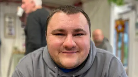 Jay Street is a 26-year-old man with short black hair. This photograph is of his head and shoulders. He's wearing a grey hoodie and is pictured in a café setting in Rhyl, with people mingling behind him.