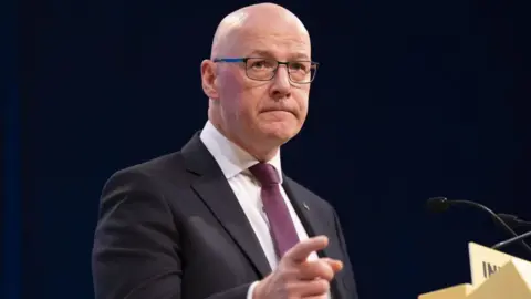 PA Media John Swinney, a bald man with glasses, points with his right hand while standing at a yellow podium. He is wearing a dark suit, white shirt and purple tie. 