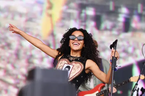 Olivia Dean performs on the Pyramid Stage at Glastonbury. She is wearing dark sunglasses and waves to the audience