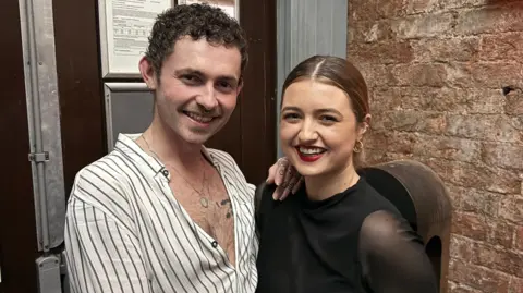 The Only Sassie has short, curly brown hair and is smiling at the camera. He is wearing a white shirt with black vertical stripes. To his right is Aisling. She is smiling at the camera and has brown hair pulled back into a ponytail.  