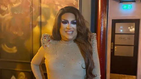 Etta Stiletta is wearing a tight fitting body suit type top in a tan colour. It has jewels and sequins sewn onto it in red and silver. The top has exaggerated pointed shoulder pads which are also bejeweled. They are wearing full drag make up and their long brown hair is pulled over their left shoulder.  