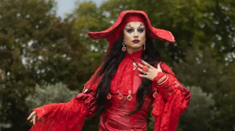 Bones wears a red gown with red jewels a large red hat. They have long black hair.