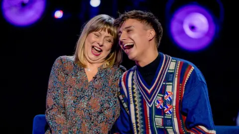 BBC/World of Wonder/Guy Levy Paul Greaves wears a blue jumper with a red, blue and white pattern. They are wearing three badges. They are sat beside their mother Diane who is wearing a patterned blouse. She has blond hair and the pair are laughing.