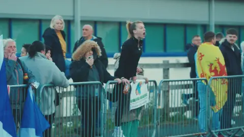 A number of protesters standing behind a barrier outside the hotel. One woman, dressed in black, is balancing on the barrier and appears to be shouting.