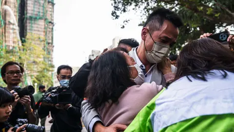 Getty Images An elderly woman, in a light purple top, cries and holds onto a young man in front of scafolded buildings. They are surrounded by journalists. 