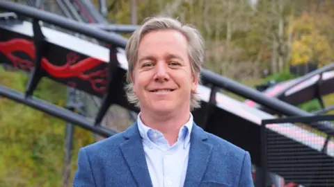 Derbyshire County Council Neil Crittenden in a blue jacket and blue shirt standing in front of a section of the Nemesis Reborn rollercoaster at Alton Towers