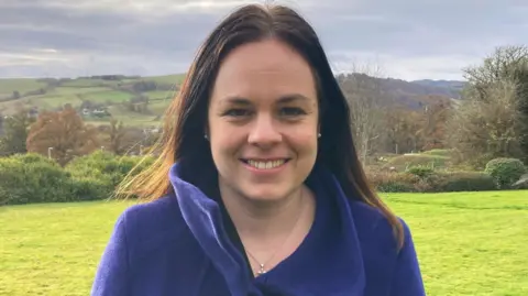 BBC Kate Forbes, a dark-haired woman in a royal blue coat stands in a lush green field with hills rolling away behind her and trees and houses in the distance.