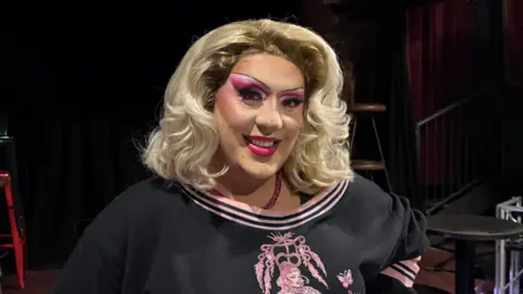 Trudy has blonde hair down to her shoulders which is shaped into curls at the end. She is wearing lots of pink eye shadow and pink lipstick along with a black and pink top. 