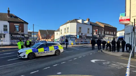 BBC/Tom Ingall A parked police car and several police officers standing in a the road. The area is cordoned off by blue and white police tape.