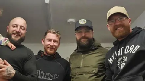 BBC Four men, three in black clothing and one wearing a green coat, are pictured shoulder to shoulder in a white room. 