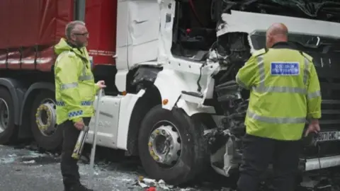 Gwent Police Investigators look at crashed lorries. They are both wearing hi-vis. The white and red lorry shows the impact of the crash.