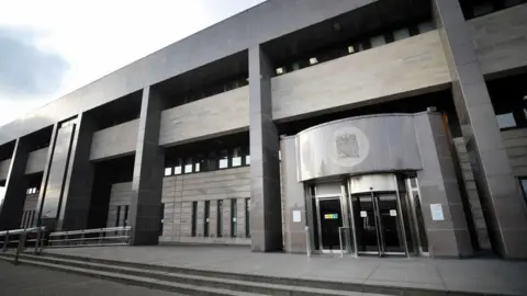 PA Media A modern grey building with a revolving door entrance with a crest above it. 
