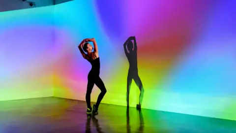 Getty Images a female dancer in a black top and leggings holds a pose while her shadow is case on a multi-coloured background on a stage