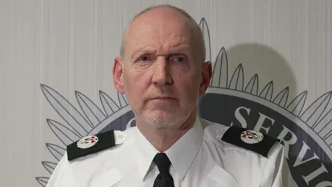 BBC A still of Davy Beck at a press conference. He is an older, bald man with a white beard. He is wearing a police uniform and has a neutral expression on his face.