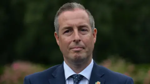 Getty Images Paul Givan stood outside with a neutral expression.   He has short, greying hair and is wearing a navy suit, a white shirt and a navy spotted tie. 