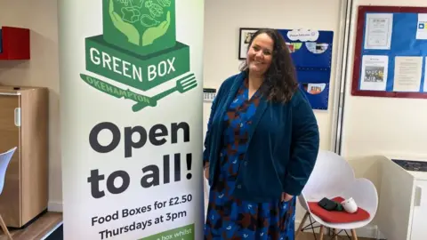 BBC Melissa Trudgill, community engagement officer for Dartmoor Multi Academy Trust is wearing a dark blue patterned dress in front of a sign which reads: Green Box, Okehampton, Open to all!
