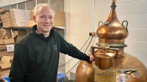 BBC Mick Skerratt in a black fleece is standing with one hand on the copper distiller which is named Isabelle. There are numerous cardboard boxes in the background and two plugs connected to a power socket in the wall.