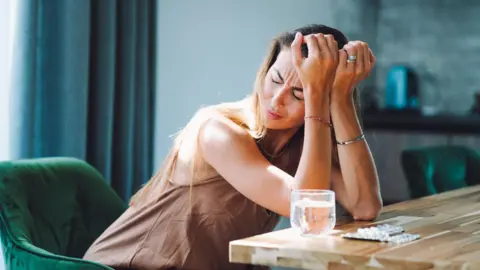 Getty Images A woman sits at a table, pressing her palms to her forehead. On the table is a glass of water and some tablets.